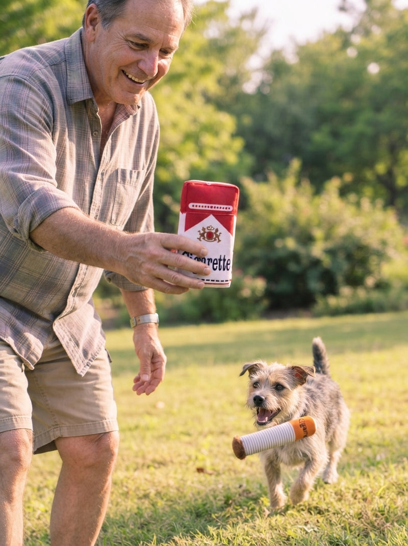 A man playing with a plush cigarette-shaped dog toy in a park with his dog nearby.